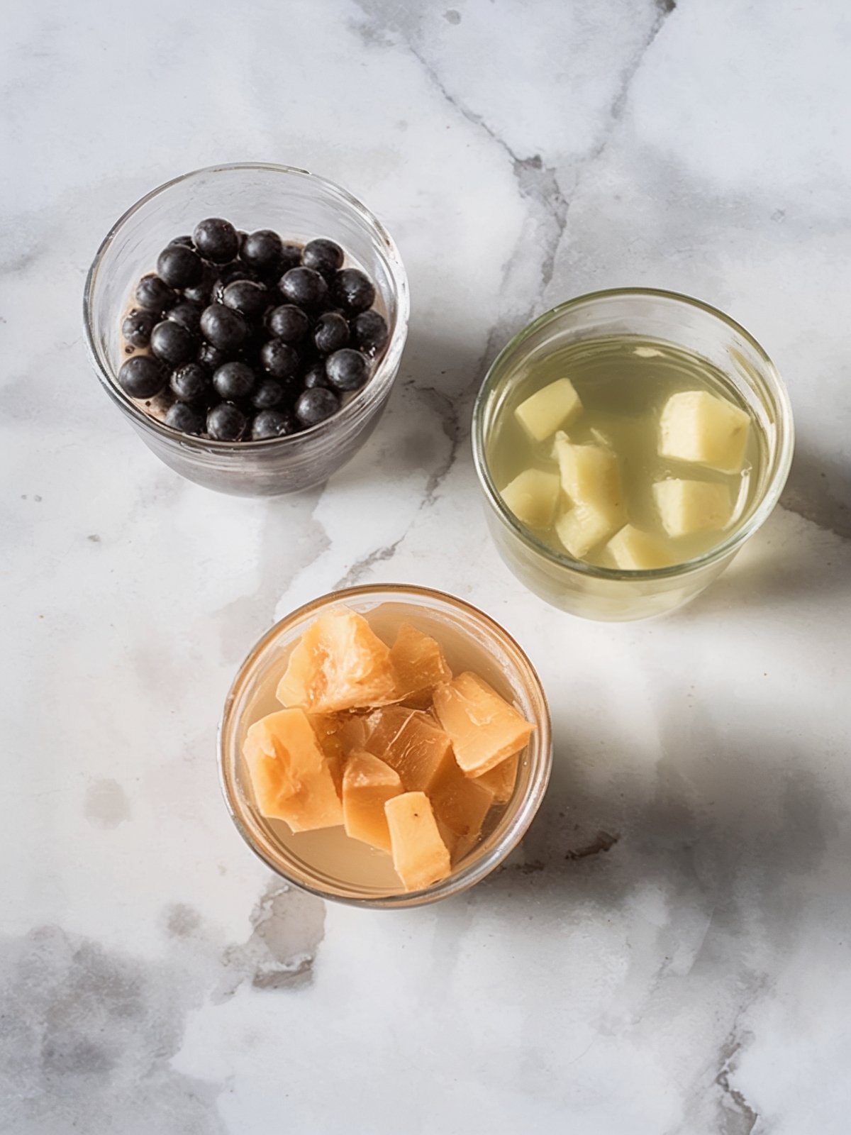 Three glass bowls with bubble tea toppings: black tapioca pearls, pale lychee jelly cubes in syrup, and brown jelly cubes on a marble countertop.