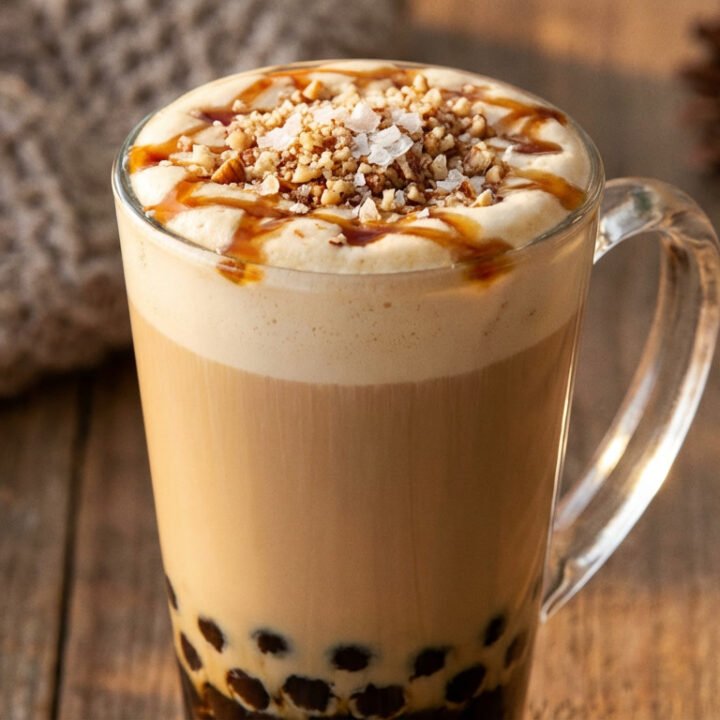 Close-up of a Salted Maple Pecan Boba Latte in a tall glass, with boba, layers of tea and milk, and topped with foam, maple drizzle, and pecans.