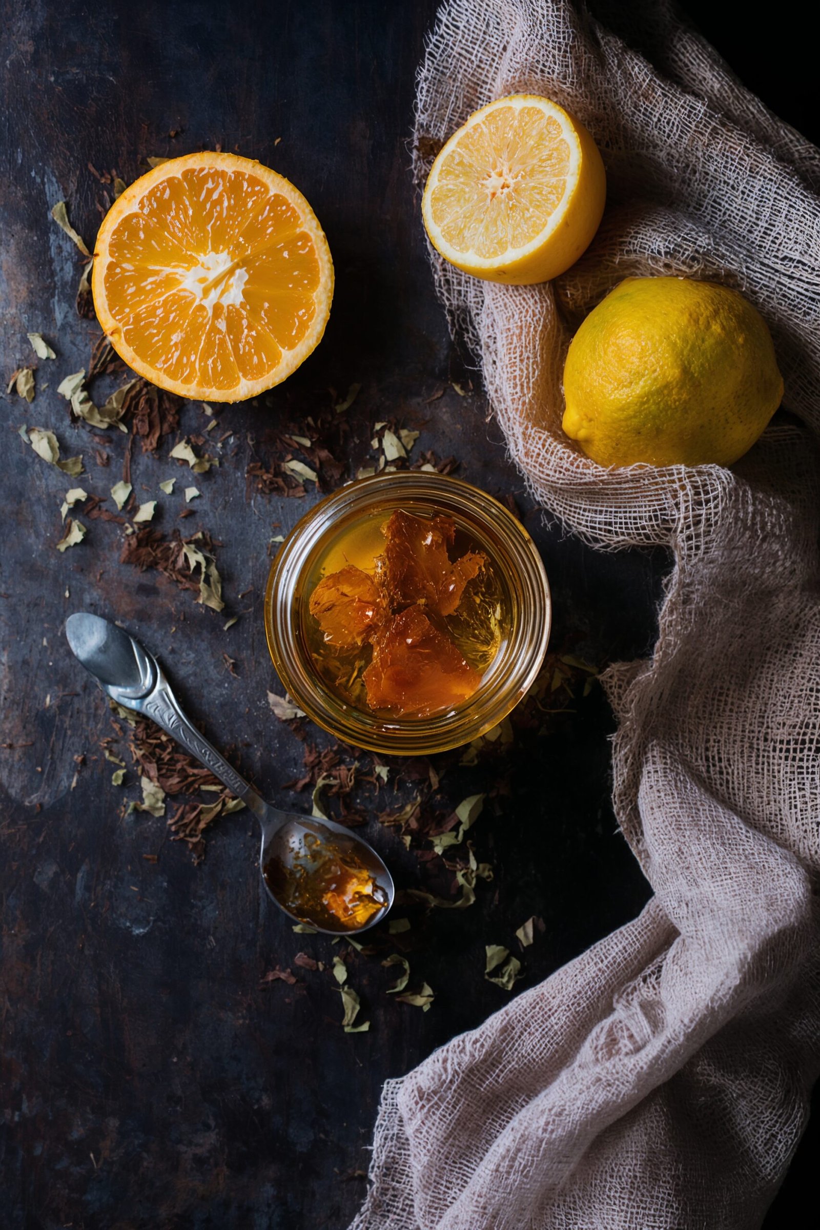 Overhead shot of raw honey in a jar, a honey-coated spoon, a halved orange, and loose jasmine tea leaves on a dark wooden table.